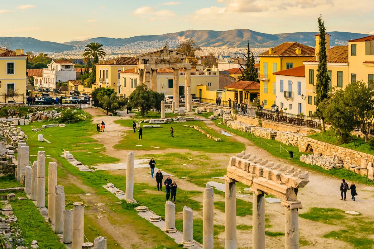 A view of the Ancient Agora in Athens, featuring scattered ruins, columns, and grassy pathways, surrounded by colorful buildings and a backdrop of distant hills.